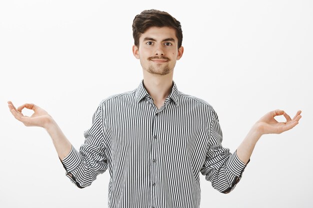 Keep mind and body in fit with yoga. Calm good-looking positive guy with beard and moustache, spreading hands in zen gesture and smiling with relaxed expression, finishing meditation over gray wall