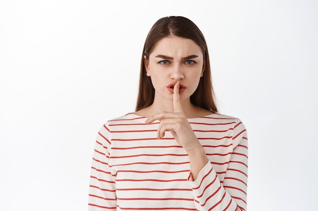 Keep quiet. Scary girl tell to shut up, shushing, hushing with angry frowning face, taboo gesture, dont talk, standing displeased against white background