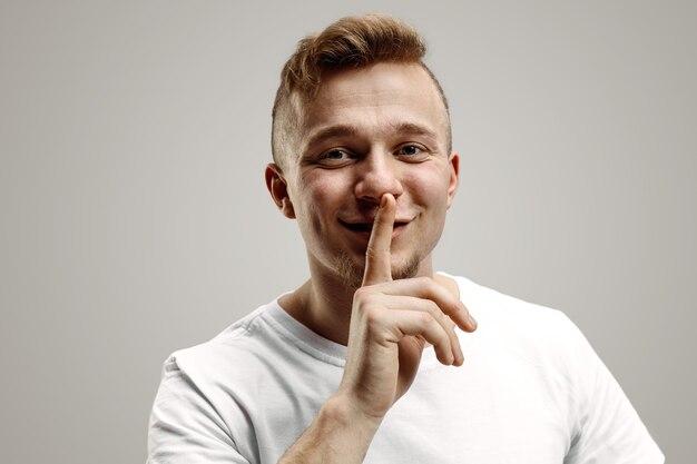 Keep silence. Handsome young man in white shirt looking at camera and holding finger on lips while standing against grey background