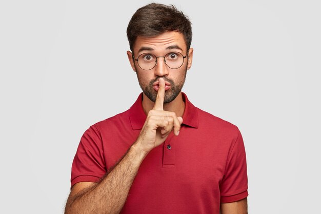 Keep voice down. Attractive surprised bearded male makes shush gesture, demnads shut up, wears casual bright red t-shirt, poses against white wall. People, hush, conspiracy, secret concept