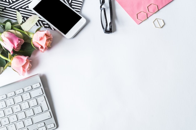 Tastiera, smartphone, occhiali e rose rosa su una superficie bianca