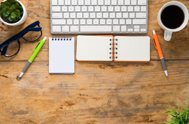 Keyboard; spiral notepad; coffee cup; eyeglasses and pen on wooden desk