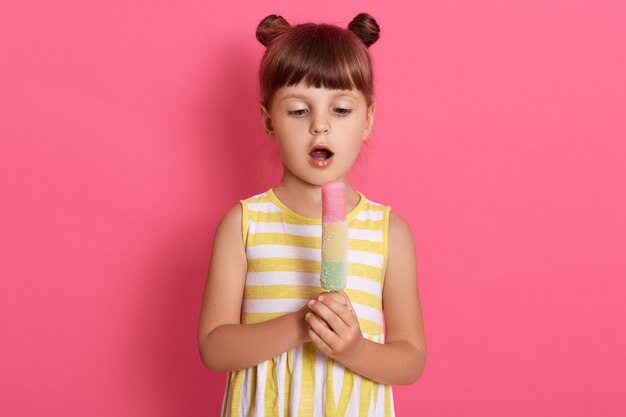 Kid eating ice cream, Funny child with two knots wearing white and yellow dress, standing with opened mouth, looking at sorbet in her hands, posing isolated on pink background.