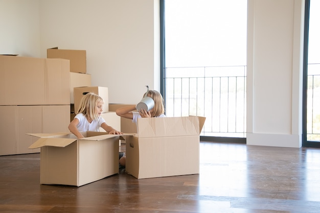 Kids having fun while unpacking things in new apartment, sitting on floor and taking objects from open cartoon boxes