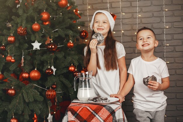 クリスマスツリーの近くに立っている子供たち。牛乳と一緒にクッキーを食べる子供たち。