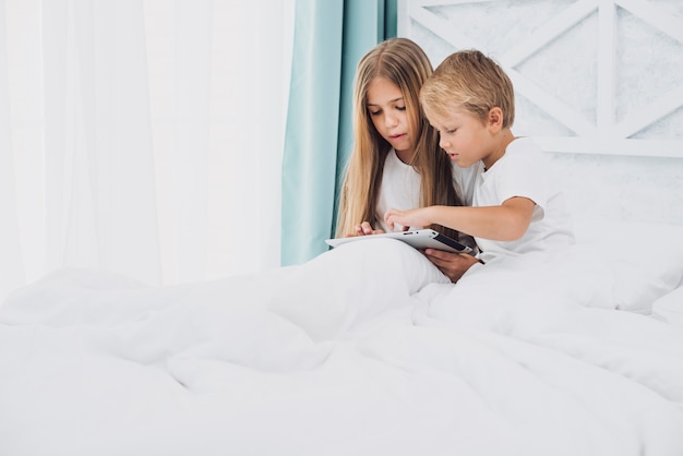 Дети, лежащие в кровати во время игры на планшете