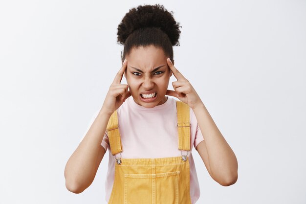 Killing you with power of mind. Portrait of angry dark-skinned female student in yellow overalls, holding index fingers on temple, staring with hate and agression over grey wall