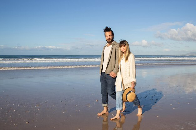 여름날 물 근처를 걷는 친절한 커플. 캐주얼 옷을 입은 수염 난 남자와 여자가 손을 잡고 해변을 산책하고 카메라를 바라보고 있습니다. 사랑, 여행, 데이트 개념