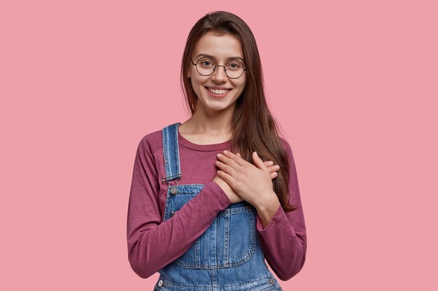 Kind European woman with pleasant smile, express favour, keeps both hands on chest, being kind hearted and honest, dressed in denim overalls