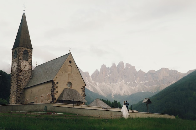Kissing wedding couple stands before stone church in mountains 