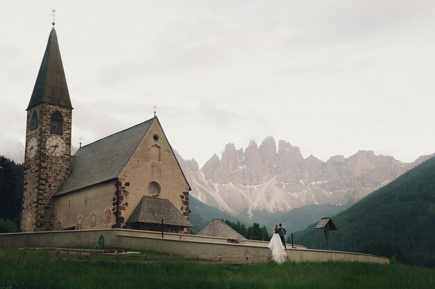 Kissing wedding couple stands before stone church in mountains 