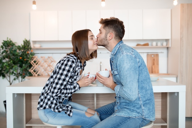 Kissing young happy man and woman in kitchen, breakfast, couple together in morning, smiling, having tea, kissing, love