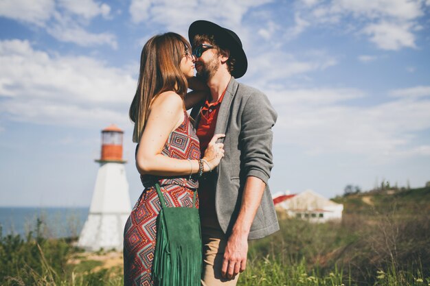 Kissing young hipster couple indie style in love walking in countryside, lighthouse on background