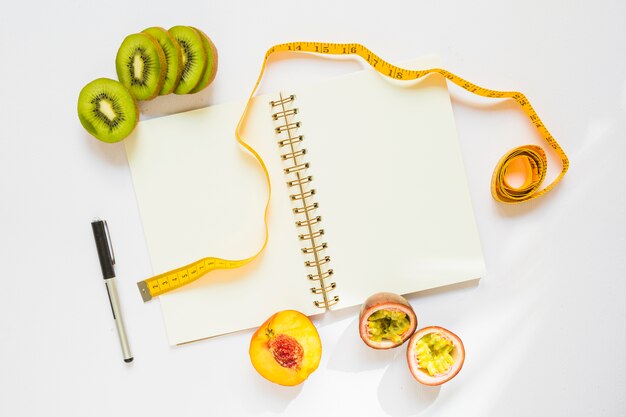 Киви ломтики; персик и страсть с измерительной лентой; ручка и спиральный ноутбук на белом фоне