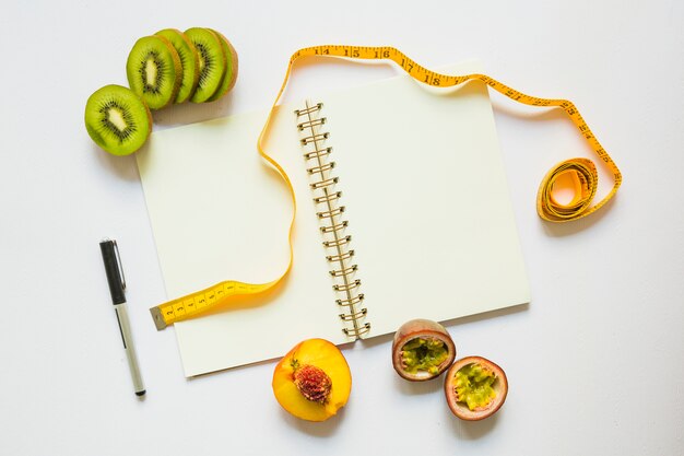 Kiwi slices; peach and passion fruits with measuring tape; pen and spiral notebook
