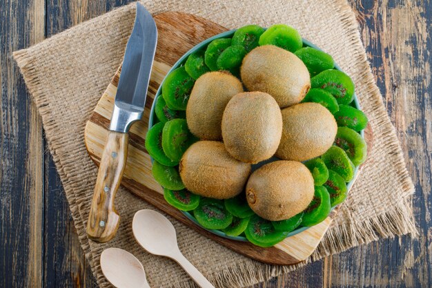 Kiwi with dried slices, cutting board, knife, spoons in a plate on wooden and piece of sack background, flat lay.