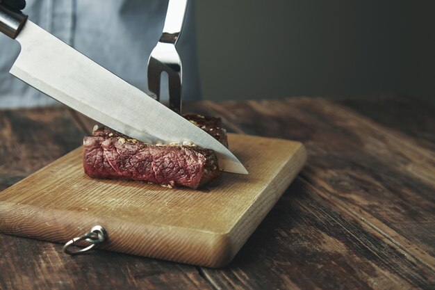 Knife cut slice of grilled meat on wooden board in front of big steel fork in steak.