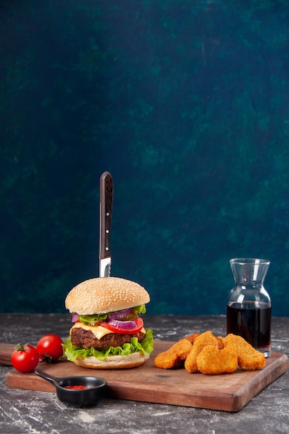 Knife in meat sandwich and chicken nuggets tomatoes with stem on wooden board sauce ketchup on dark blue surface