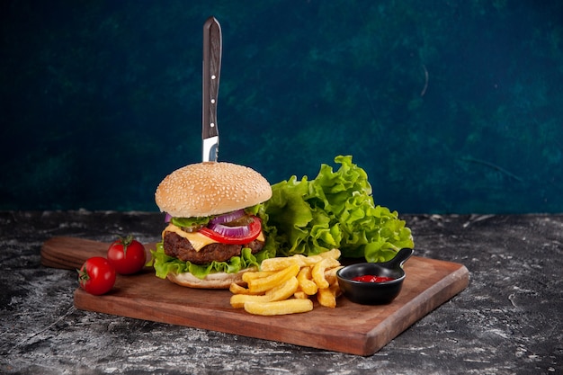 Knife in meat sandwich and fries tomatoes with stem pepper on wooden board ketchup on dark blue surface