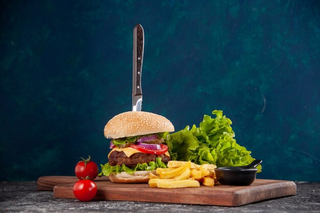 Knife in meat sandwich and fries tomatoes with stem on wooden board ketchup on dark blue surface