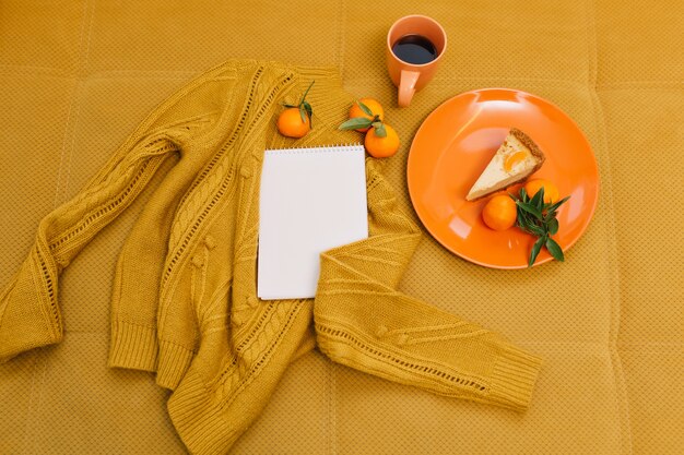 Knitted sweater, cup of coffee, cheesecake, mandarines, notebook on orange table