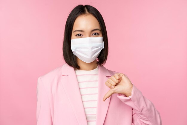 医療用フェイスマスクとスーツを着た韓国人ビジネスウーマンは、ピンクの背景の上に立っているジェスチャーを嫌いまたは不承認の親指を下に示しています