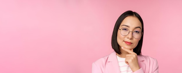 Korean businesswoman thinking wearing glasses looking thoughtful at camera making decision standing over pink background