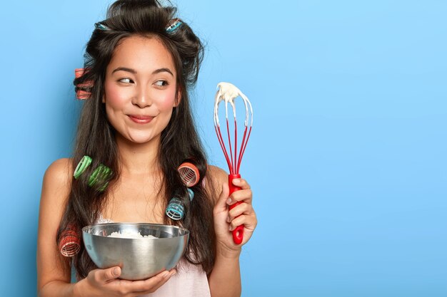 Korean housewife with hair curlers, busy cooking and preparing for birthday party, whisps cream with hand whisk