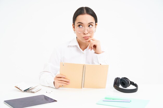 Korean woman hides her face behind notebook peeks at camera sits at desk with headphones and tablet