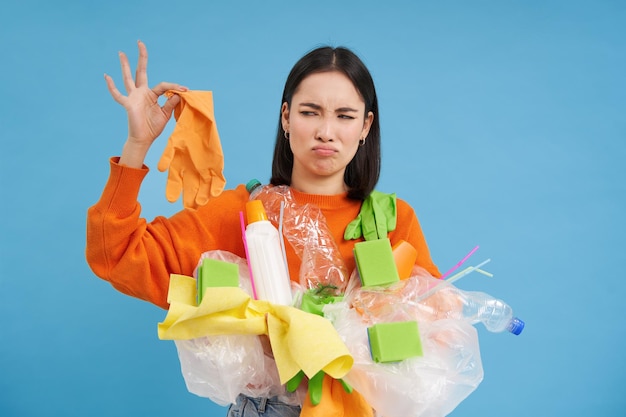 Бесплатное фото Кореянка смотрит на вонючую латексную перчатку, держит пластиковый мусор, убирает дом и перерабатывает синий бэкгро