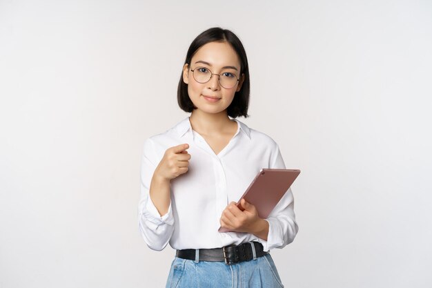 Korean woman office worker manager in glasses holding working tablet and pointing at you choosing recruiting standing over white background