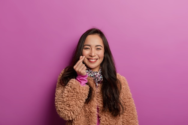 Korean woman shows mini heart sign, smiles tenderly, expresses affection and love during photoshoot