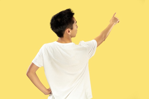 黄色のスタジオの背景に韓国の若い男の半身像。白いシャツの男性モデル。広告の場所を指しています。人間の感情、顔の表情の概念。トレンディな色。
