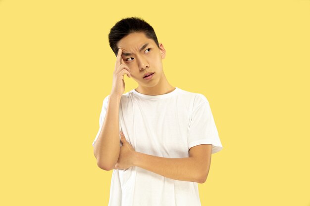 Korean young man's half-length portrait on yellow studio background. Male model in white shirt. Serious thinking or thoughtful. Concept of human emotions, facial expression. Front view. Trendy colors.