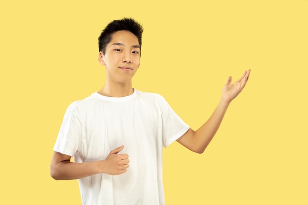 Поясной портрет корейского молодого человека на желтом студийном фоне. Мужская модель в белой рубашке. Показывает что-то. Понятие о человеческих эмоциях, выражении лица. Передний план. Модные цвета.
