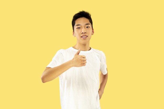 노란색 스튜디오 배경에 한국 젊은이의 절반 길이 초상화. 흰 셔츠에 남성 모델. 웃고 OK의 표시를 보여줍니다. 인간의 감정, 표정의 개념.