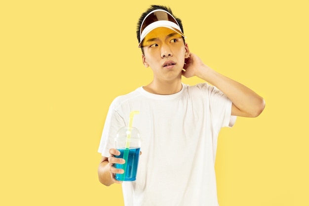 Поясной портрет корейского молодого человека на желтом студийном фоне. Модель-мужчина в белой рубашке и желтой кепке. Питьевой коктейль. Понятие человеческих эмоций, выражения, лета, отпуска, выходных.