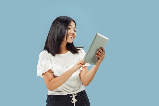 Поясной портрет корейской молодой женщины на синем пространстве. Женская модель в белой рубашке. Используя планшет и улыбаясь.