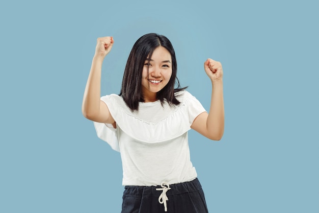 Korean young woman's half-length portrait. Female model in white shirt. Celebrating like a winner, looks happy. Concept of human emotions, facial expression. Front view.