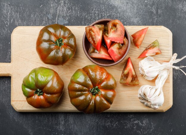 Kumato tomatoes with slices, garlic bulbs on grunge and cutting board wall, top view.