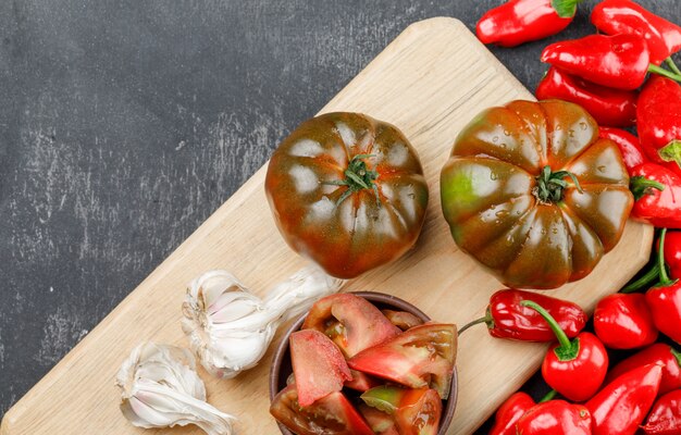 Kumato tomatoes with slices, red peppers, garlic bulbs on grey and cutting board wall, flat lay.