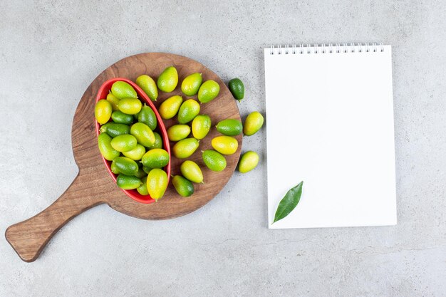 Kumquats piled in and next to a bowl on a wooden board, next to a leaf-adorned notebook on marble surface