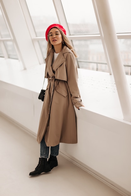 Lady in beige midi trench and red beret with crossbody bag, posing leaning on large white window