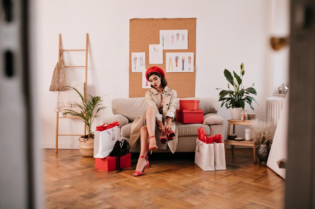 Lady in beige modern coat and red hat looks at her shoes Cute young woman in fashionable heels is trying on her purchase