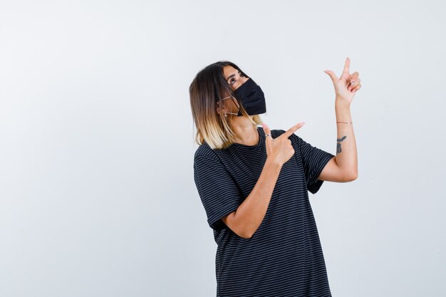 Lady in black dress, medical mask pointing up and looking hopeful , front view.