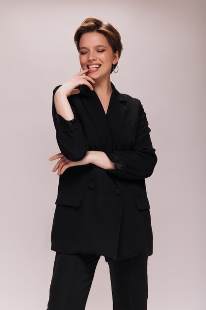 Lady in black suit happily posing on white background. Cheerful woman in dark jacket and pants smiling on isolated
