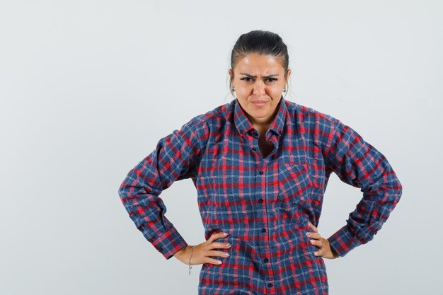 Lady in casual shirt holding hands on waist and looking spiteful , front view.