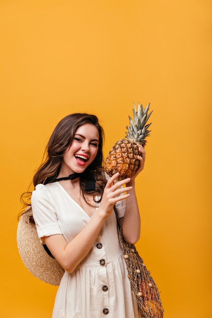 Lady in cotton dress is holding pineapple. Woman with wavy hair posing with shopping bag on orange background.