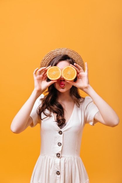 Lady covers her eyes with oranges. Woman in straw hat and white outfit blows kiss.