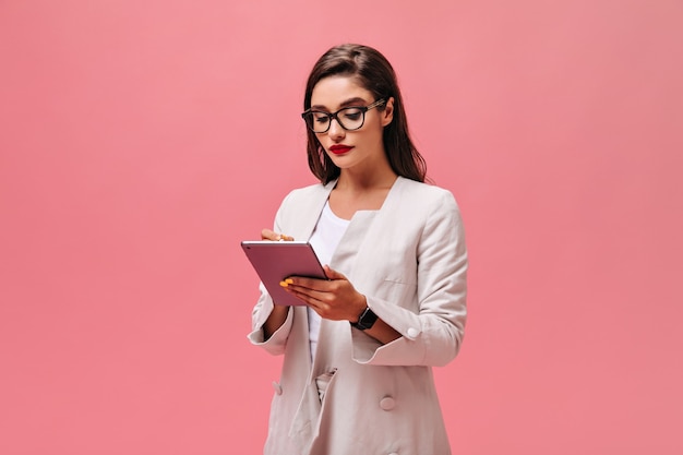 Free photo lady in eyeglasses and jacket holds computer tablet on pink background.  business woman with red lips in bright clothes writes something.